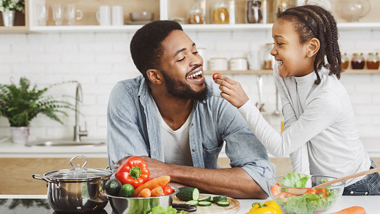 young child feeding a vegetable to an adult in the kitchen preparing a healthy meal